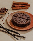 Chocolate cake with 'Fudgy' branding on a plate, surrounded by vanilla beans, cinnamon sticks, and cacao nibs.