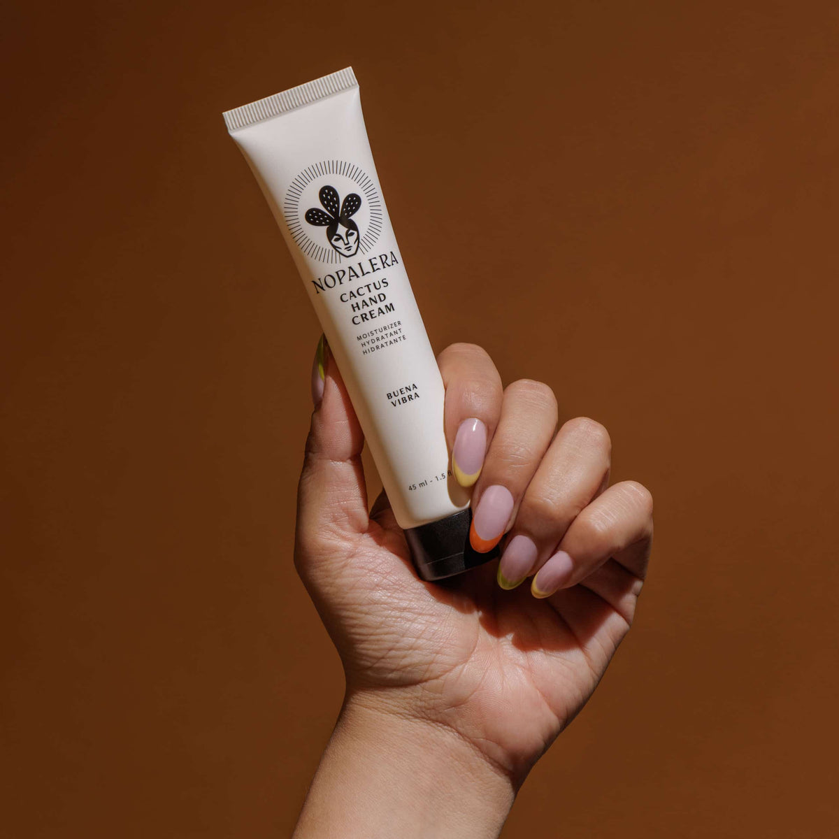 A hand with pastel multicolor nails holds a squeeze tube of Nopalera Cactus Hand Cream against a solid warm brown background.