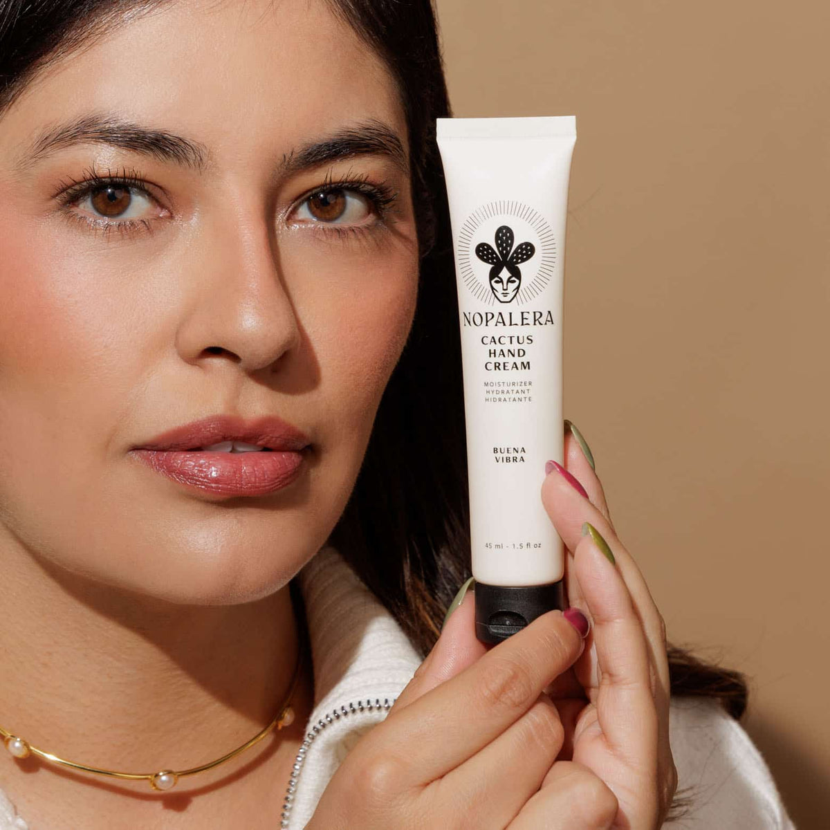 A close-up lifestyle shot of a woman holding a white tube of Nopalera Cactus Hand Cream Buena Vibra against a warm beige background.