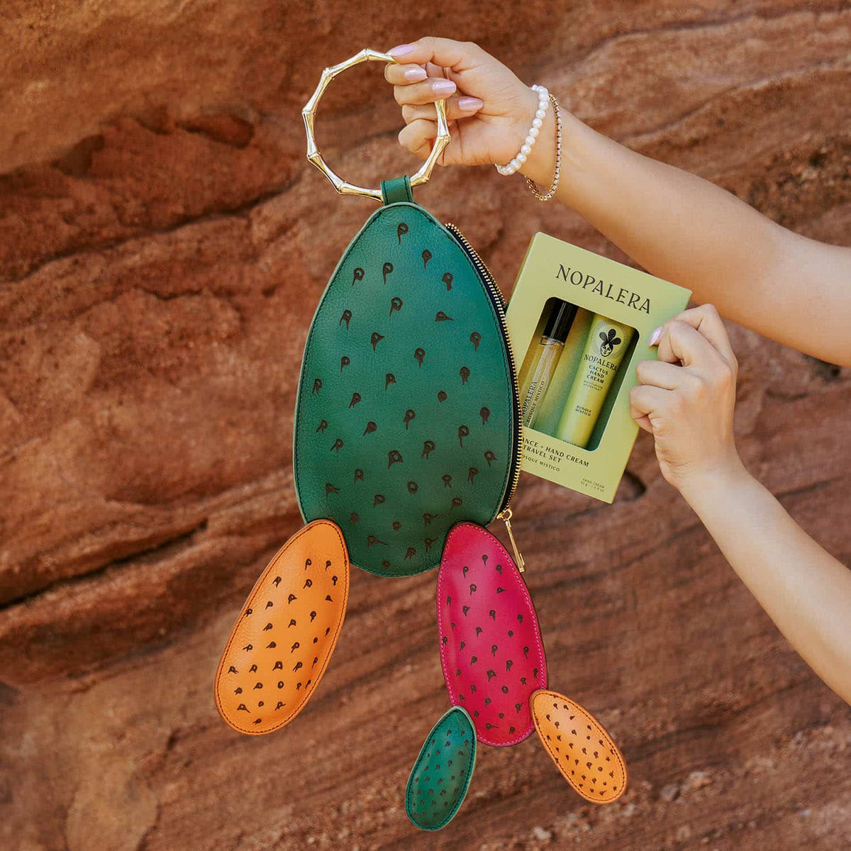 Carlota Bag in green with colorful cactus-shaped pouches, held alongside a Nopalera fragrance set against a desert rock background.