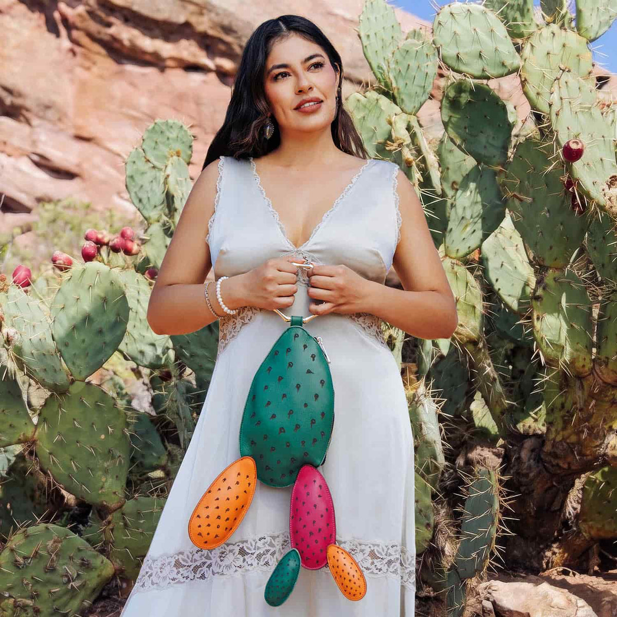 Woman in a white dress holding the colorful Carlota Bag while standing among prickly pear cacti in a desert landscape.