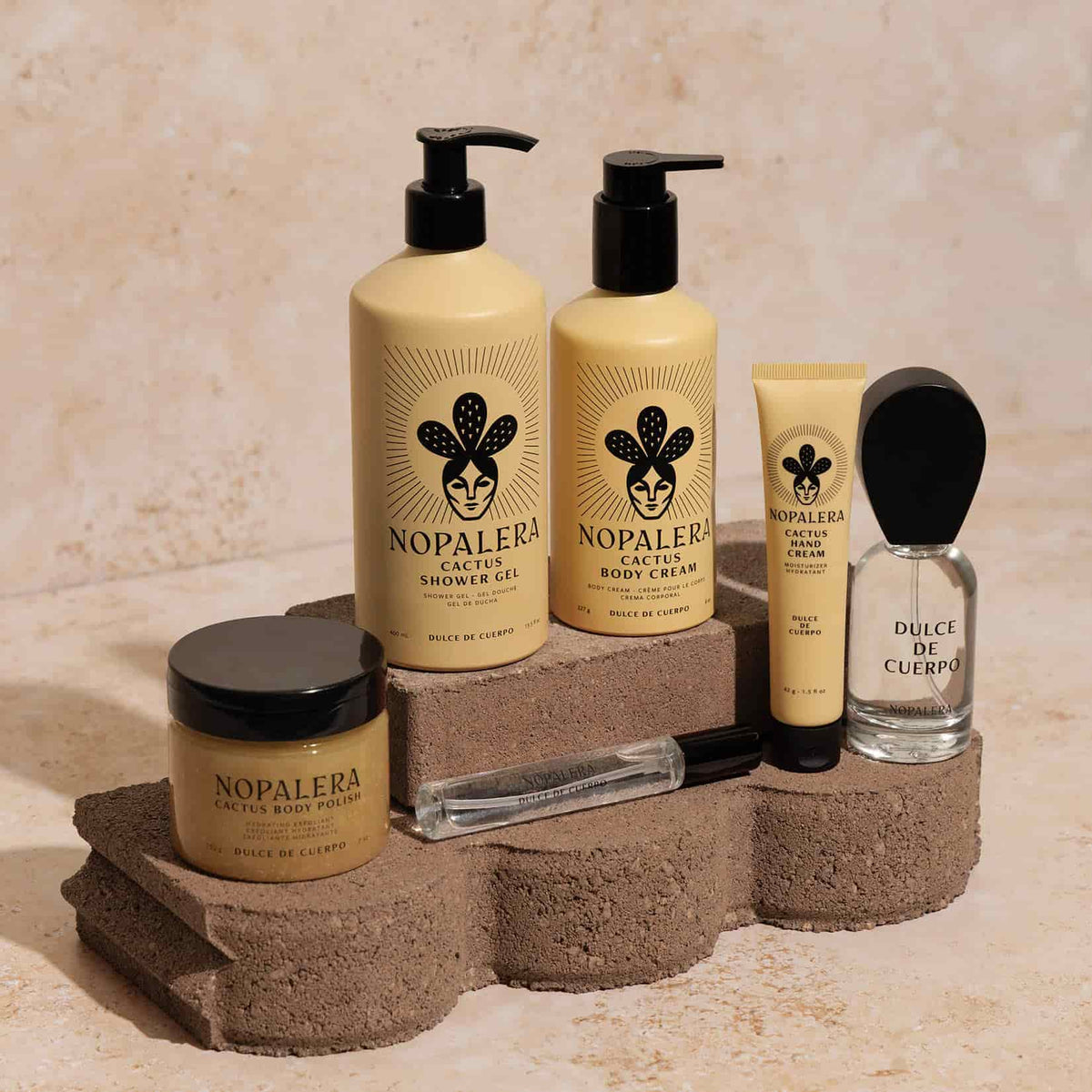 A set of Nopalera Dulce de Cuerpo skincare and fragrance products arranged on rustic stone blocks against a textured beige wall. The products include a cactus body polish jar,  shower gel, body cream, a hand cream tube, a clear perfume bottle labeled Dulce de Cuerpo, and a travel spray.