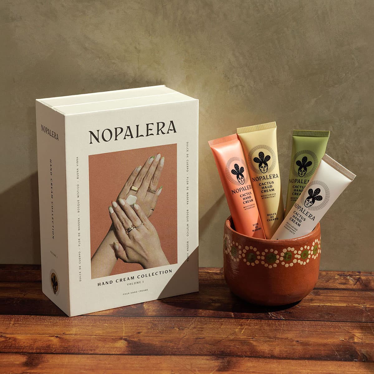 Nopalera Hand Cream Collection gift box beside four Cactus Hand Cream tubes in Flor de Madera, Dulce de Cuerpo, Bosque Místico, and Buena Vibra scents, arranged in a painted terracotta pot on a wooden table.