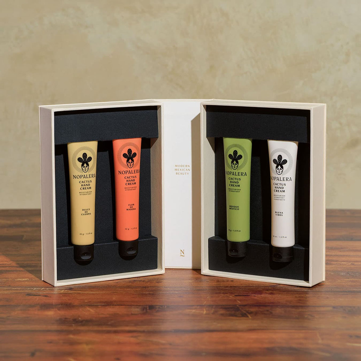 Open Nopalera Hand Cream Collection gift box revealing four Cactus Hand Cream tubes in Flor de Madera, Dulce de Cuerpo, Bosque Místico, and Buena Vibra scents, set in a black insert on a wooden table.