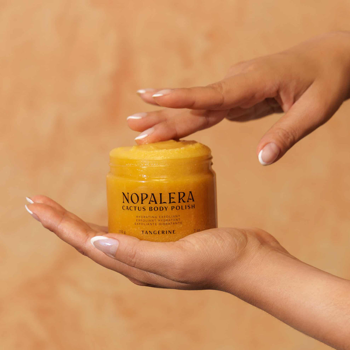Hand holding a jar of Nopalera Cactus Body Polish Tangerine against a warm-toned background