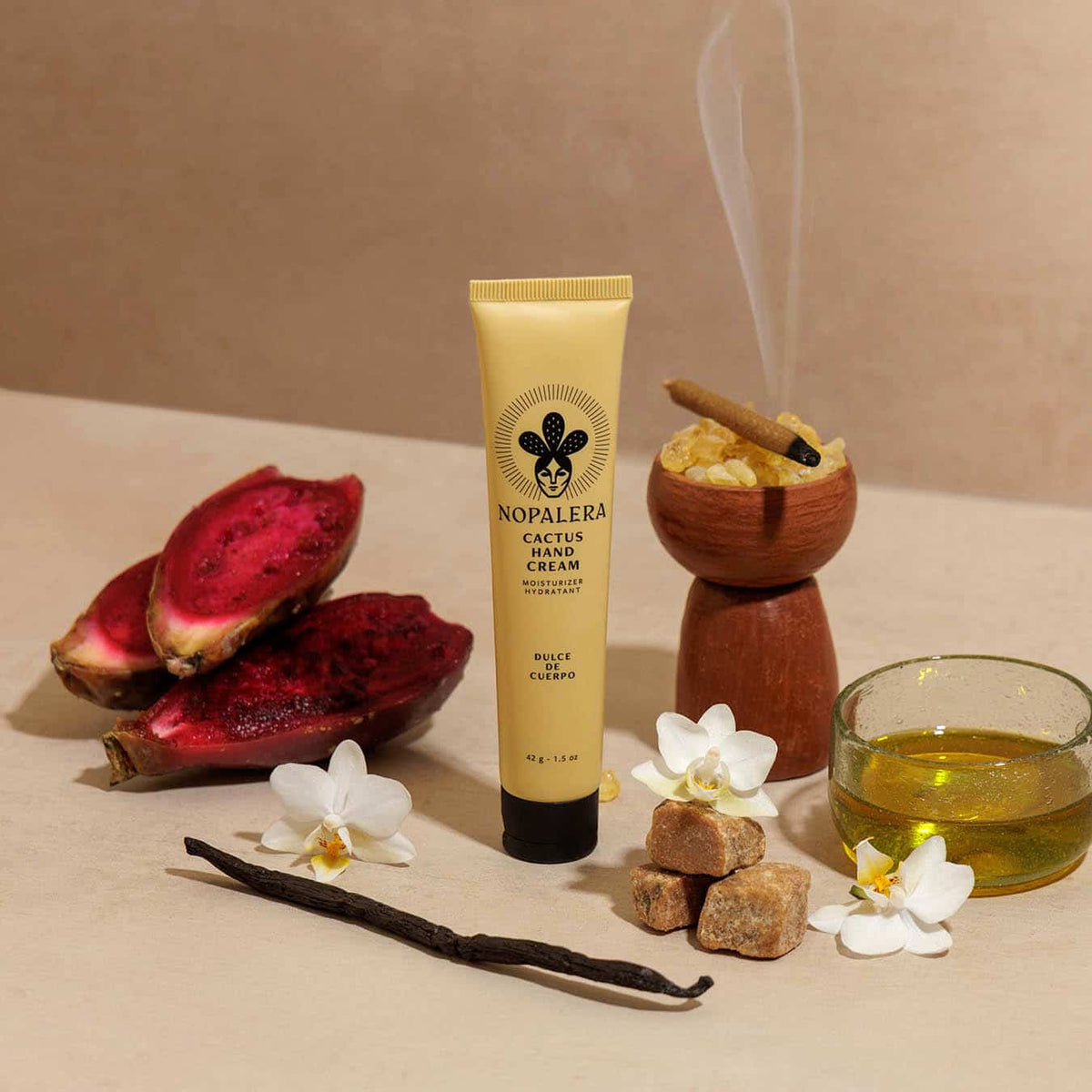 A tube of Nopalera Cactus Hand Cream stands on a beige surface surrounded by natural ingredients and aromas, including sliced red nopal, white orchids, brown sugar cubes, a vanilla bean, a glass of yellow oil, and a wooden bowl with a smoking herb.