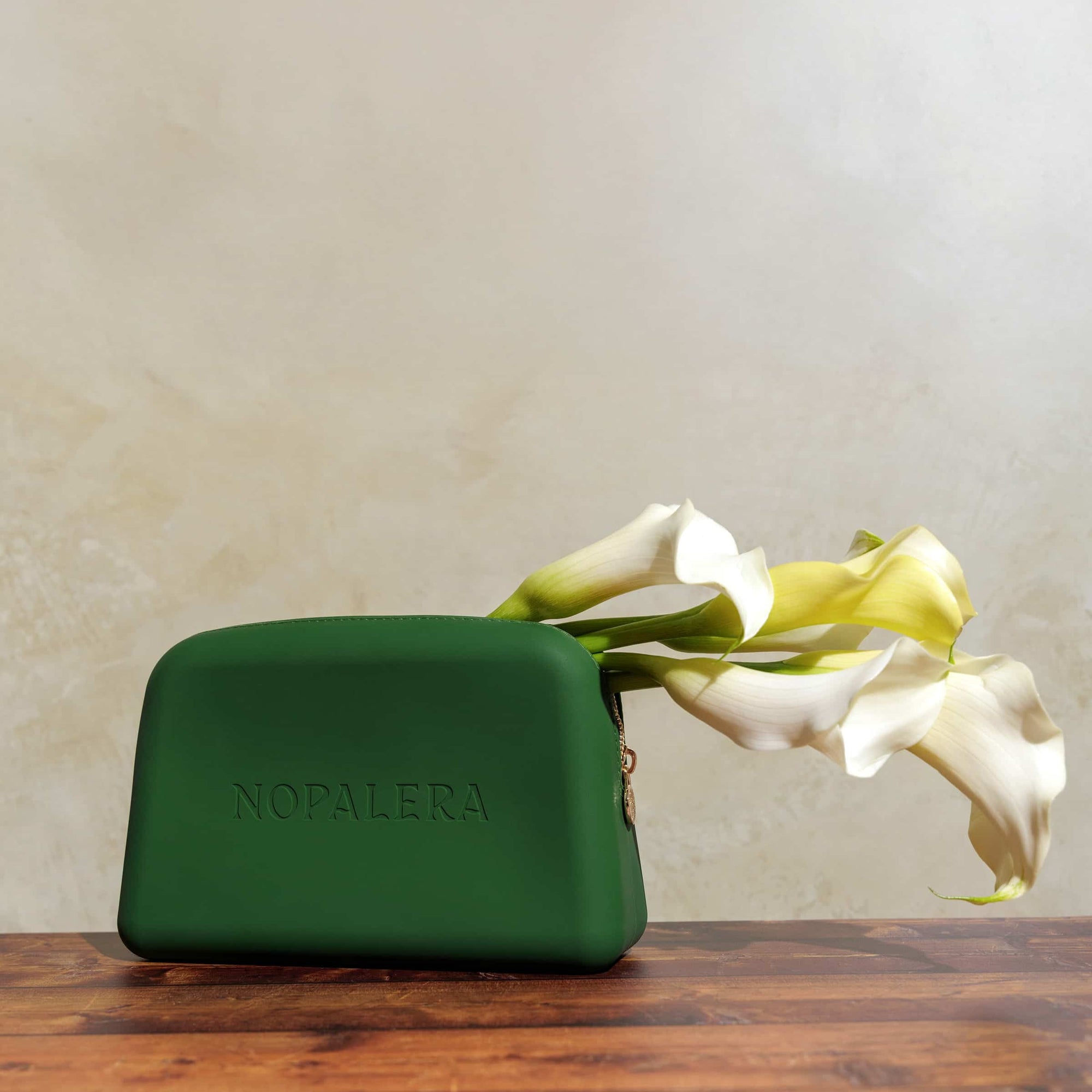 Green nopalera.sbssmetic pouch with gold zipper and embossed logo on a neutral background and flowers