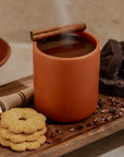 Nopalera Ceramic mug/candle with steaming hot chocolate, cinnamon stick, cookies, and chocolate on a wooden surface.